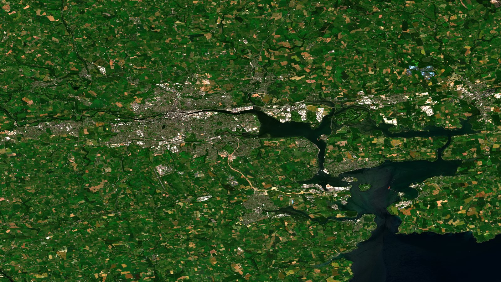 Escena Sentinel-2 en color real de Cork y su puerto. La trama urbana aparece en grises y la rodea un denso mosaico de parcelas agrícolas en verde intenso y color paja, separadas por setos. A la derecha, el río Lee se ensancha en Lough Mahon y desemboca en el puerto de Cork.