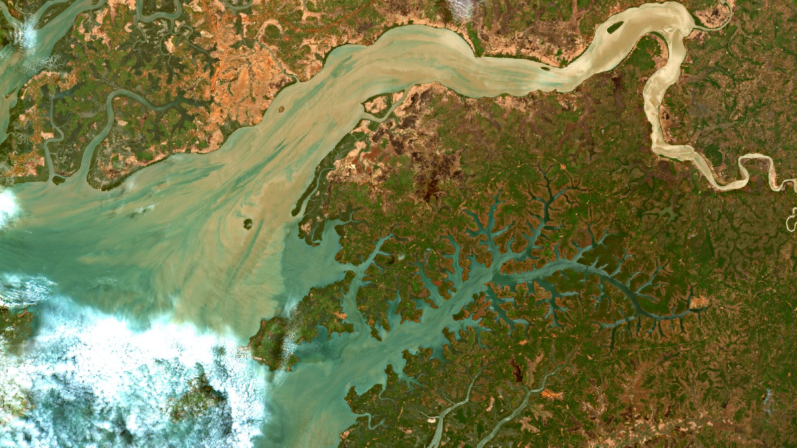 Escena Sentinel-2 en color real del oeste de Guinea-Bisáu. Arriba, el Canal do Geba serpentea de este a oeste como una cinta ancha de agua pálida cargada de sedimento. Hacia el centro y el oeste del encuadre esa agua clara se abre en abanico sobre el Atlántico. Abajo a la derecha, la red ramificada de rías del Rio Grande de Buba, con el manglar en verde oscuro moteado de pardo entre canales de agua turquesa. El interior, al este, es un mosaico de verdes oscuros y tierras rojizas de laterita. Un banco de nubes blancas ocupa la esquina inferior izquierda, sobre el mar.