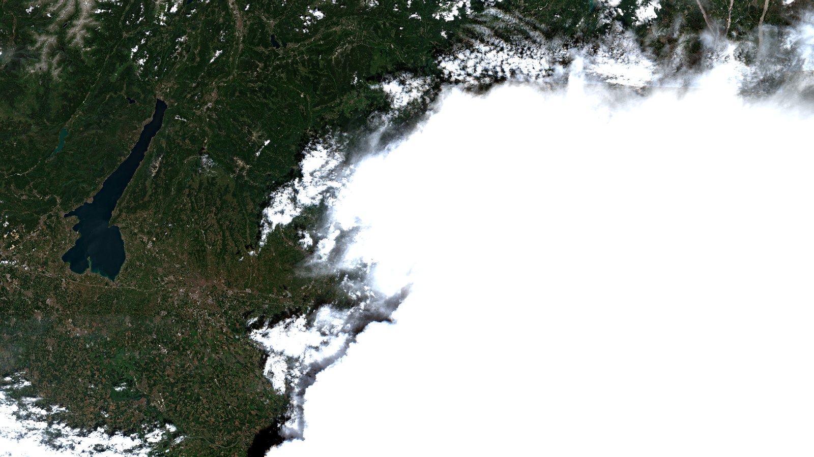 Escena Sentinel-2 del norte de Italia el 21 de julio de 2026, en color real. La mitad derecha del encuadre la ocupa un enorme yunque de tormenta, una masa de nube blanca y compacta de bordes redondeados. A su izquierda, una franja gris oscura pegada al contorno de la nube marca la sombra proyectada sobre el terreno. La mitad izquierda muestra la llanura del Po en tonos verdes y pardos, el lago de Garda como una mancha azul muy oscura alargada y, arriba, los relieves alpinos con algunas cumbres claras.